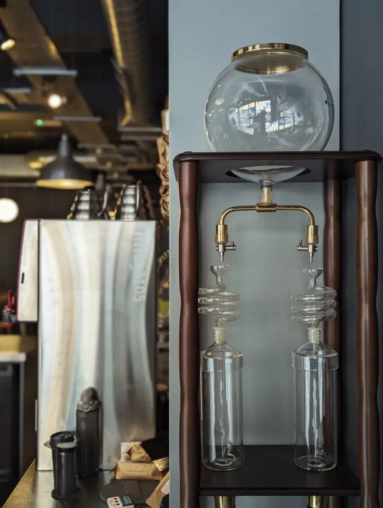 A cold brew coffee tower with glass globes, spiral glass tubes, and brass fittings on a wooden stand in a cafe setting.