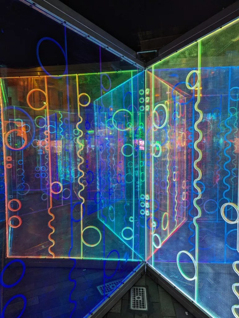 Neon rainbow circles and wavy lines glowing on glass panels, creating a reflective light installation outdoors at night.