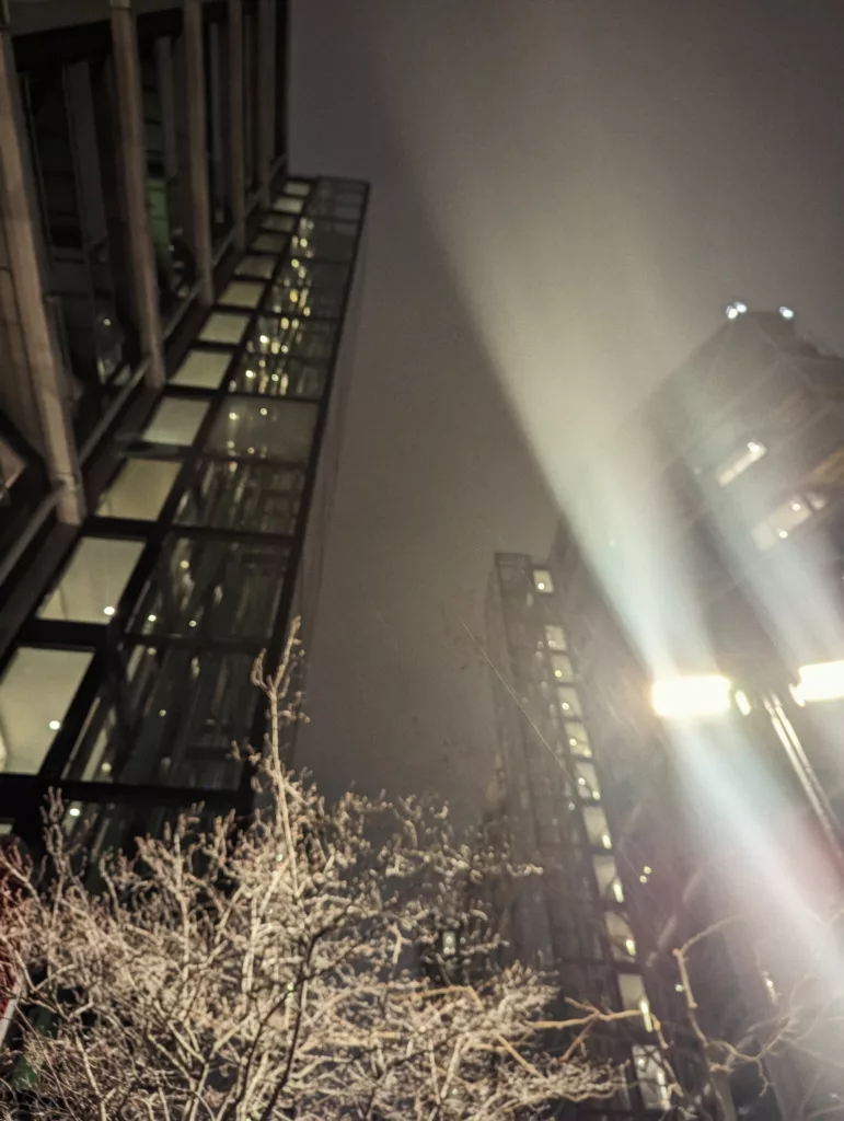 A low-angle night view of glass skyscrapers with a bright light beam cutting through misty air above bare winter branches.