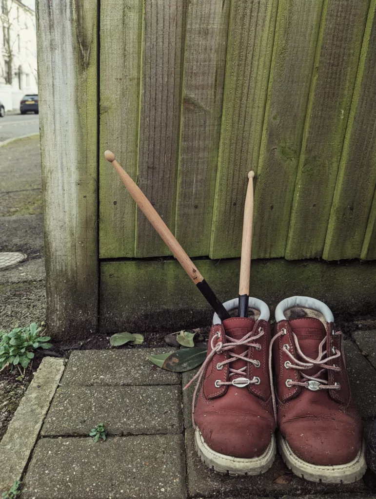 Two wooden drumsticks tucked into a maroon boot on a stone patio beside a weathered green wooden fence.