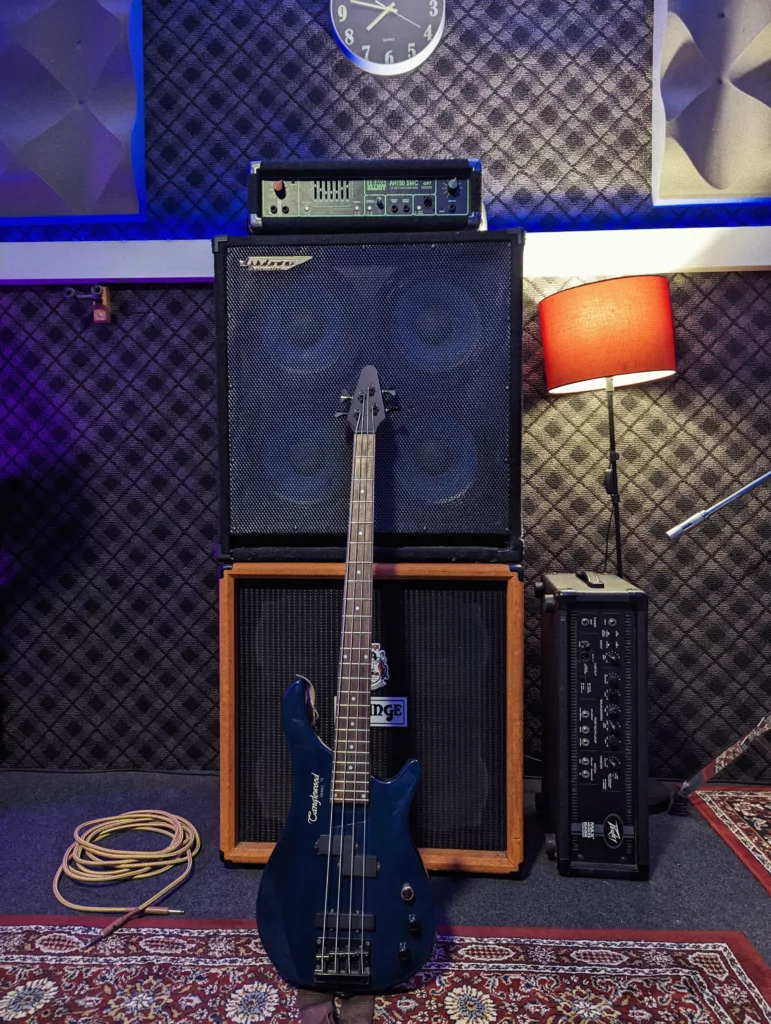 Blue Tanglewood bass guitar leaning against a stack of Orange and Trace Elliot amplifiers in a studio with soundproofed walls and a red lamp.