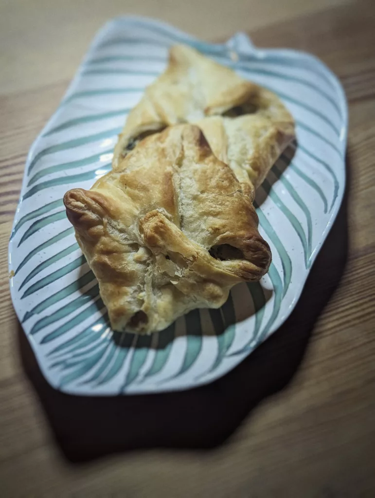 Two golden-brown puff pastry parcels on a ceramic plate with a teal palm leaf design, resting on a wooden table.