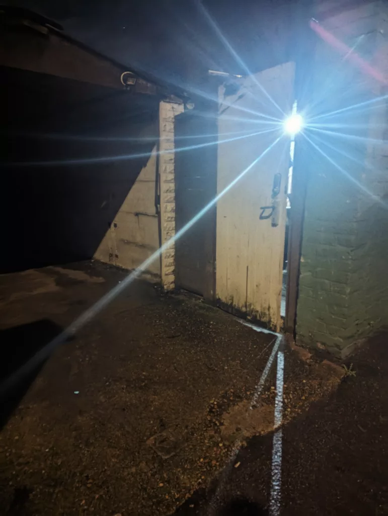 A bright white security light creates a starburst lens flare as it shines through an open white wooden door at night.