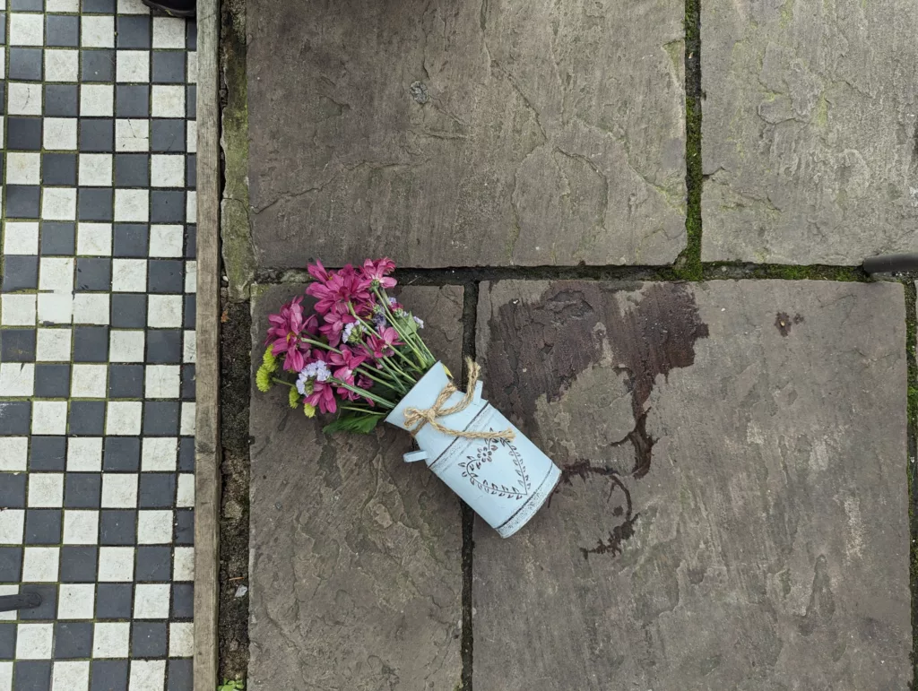 A tipped-over light blue jug of pink flowers on stone paving beside a wet spill and a black and white checkered tile border.