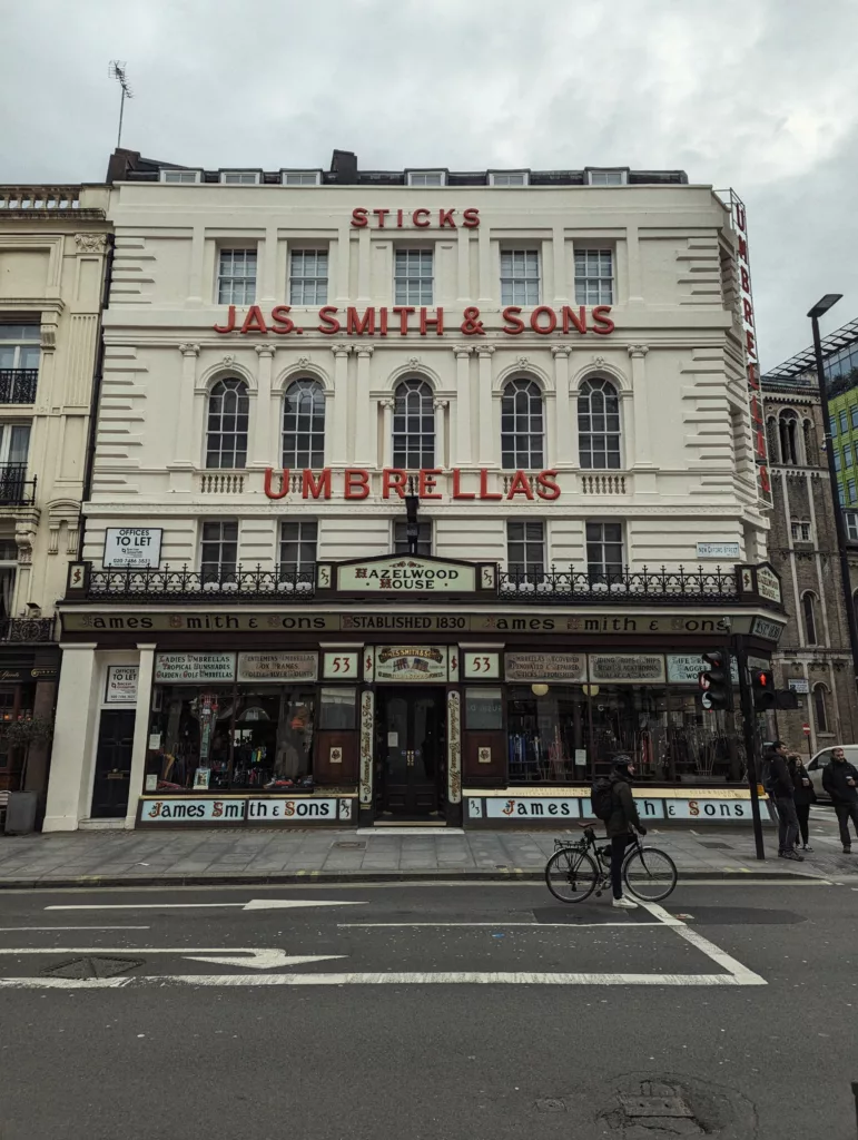 The white Victorian facade of James Smith & Sons umbrella shop in London, featuring bold red signage and a cyclist in the foreground.