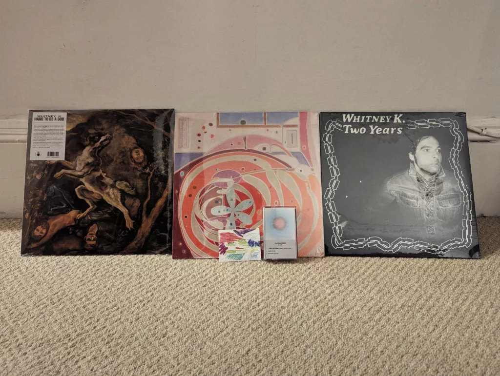 Whitney K vinyl records for Hard to be a God and Two Years, plus a cassette tape, leaning against a white wall on a beige carpet.