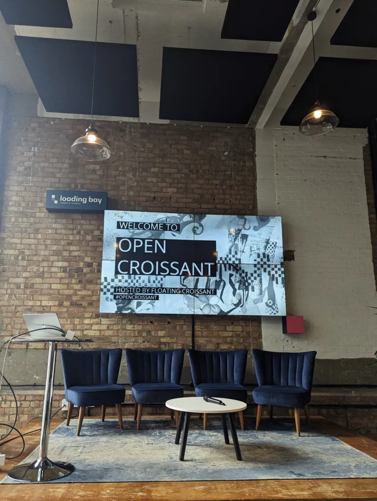 Four blue velvet chairs and a coffee table on a rug before a screen reading "Welcome to Open Croissant" on an exposed brick wall.