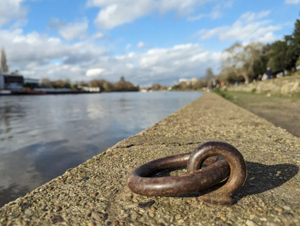A rusty iron mooring ring on a concrete riverwall, looking out over a calm river under a bright blue sky with white clouds.
