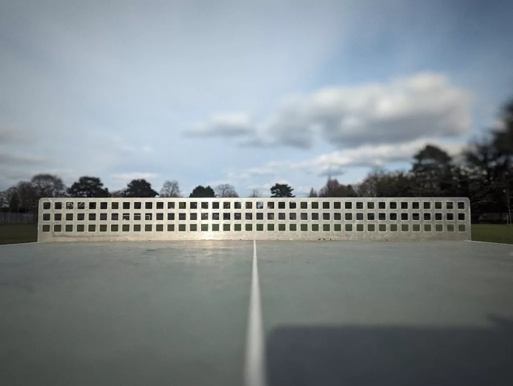 Low-angle perspective looking across an outdoor table tennis table with a metal net toward a park and cloudy blue sky.