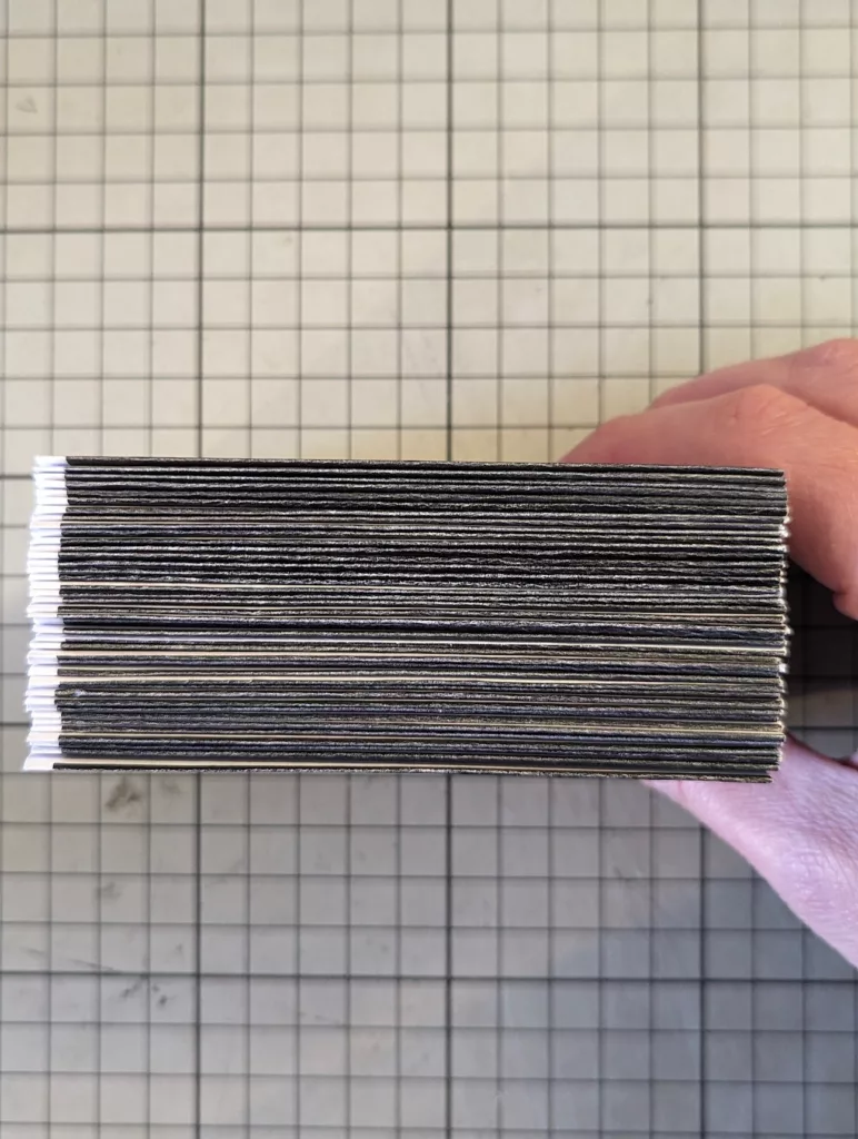 A hand holding a thick stack of black-edged cards from above, resting on a grey gridded cutting mat.