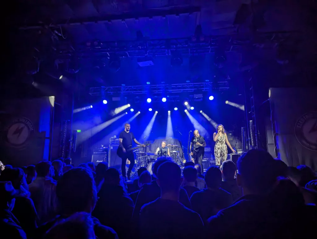 A four-piece band performs on a stage under dramatic blue spotlights, viewed from the back of a crowd in a dark music venue.