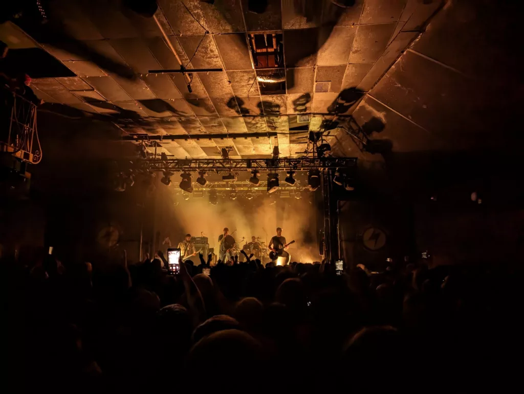 A silhouetted band performs on a smoky stage under warm amber spotlights in a dark venue with a tiled ceiling and a crowded audience.