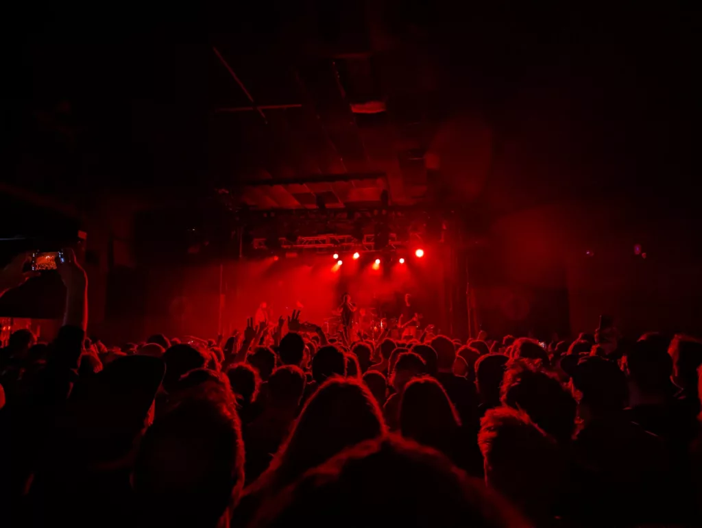 Silhouetted crowd in a dark concert venue facing a stage illuminated by intense red spotlights.