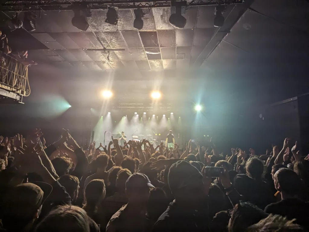 Crowds raise their hands in a dark, hazy concert venue as a band performs on stage under bright yellow and white spotlights.