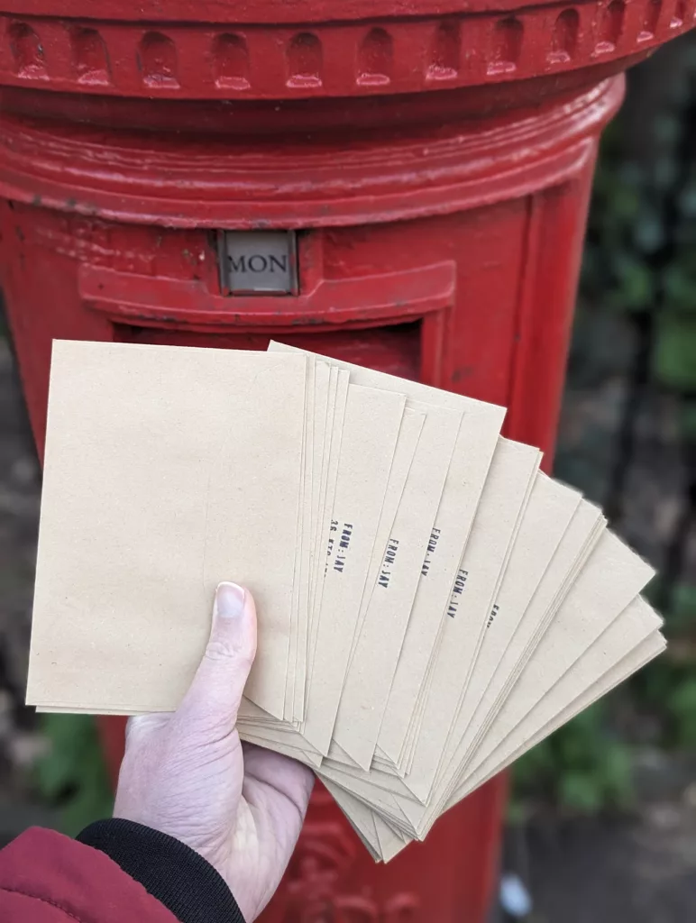 A hand holding a fanned-out stack of brown kraft envelopes stamped FROM: JAY in front of a red British pillar post box.