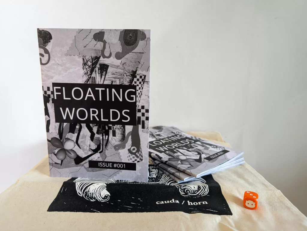Monochrome zine Floating Worlds Issue #001 with an abstract collage cover stands on a tote bag beside a stack of copies and a small orange die.
