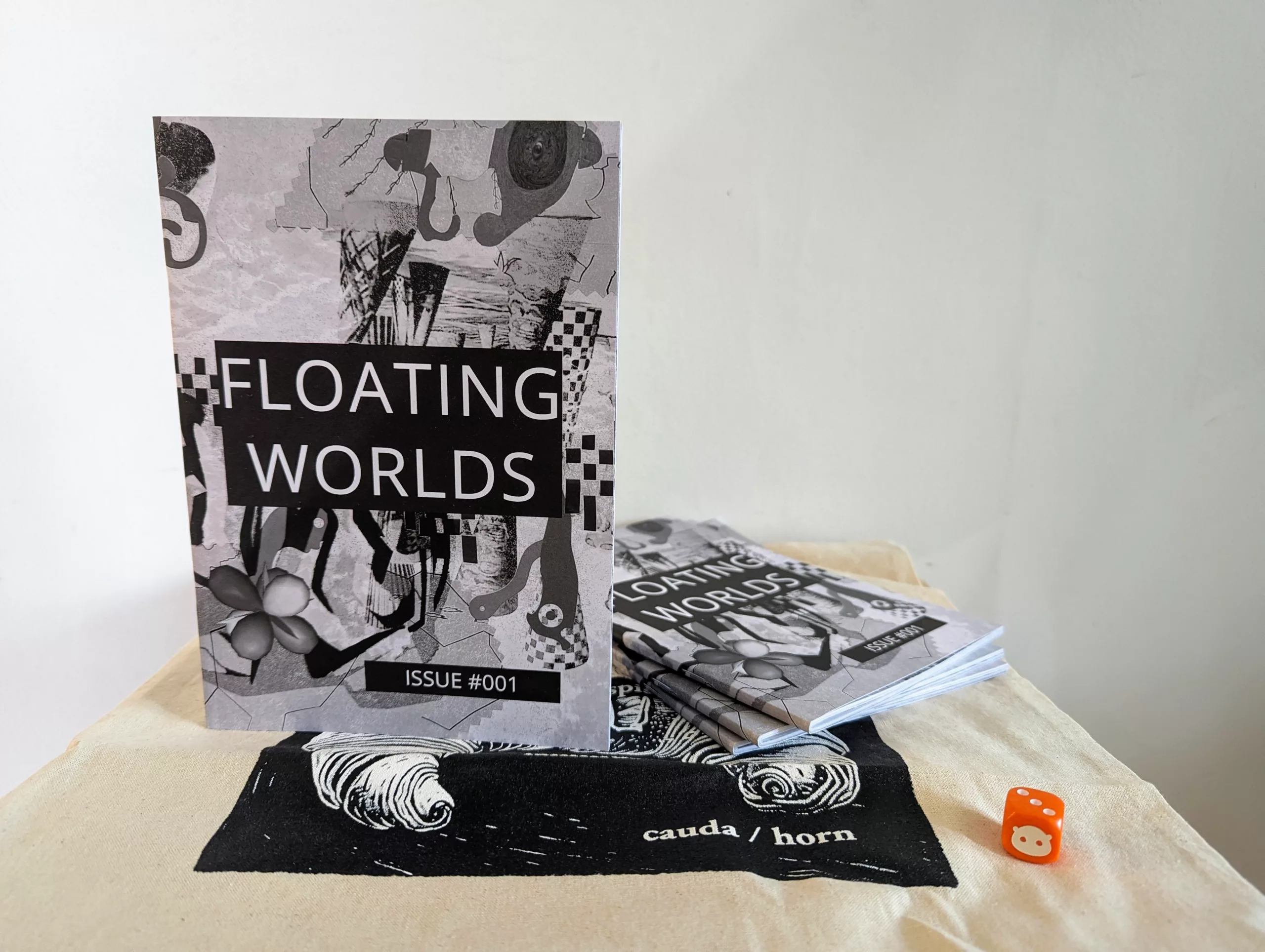 Monochrome zine Floating Worlds Issue #001 with an abstract collage cover stands on a tote bag beside a stack of copies and a small orange die.