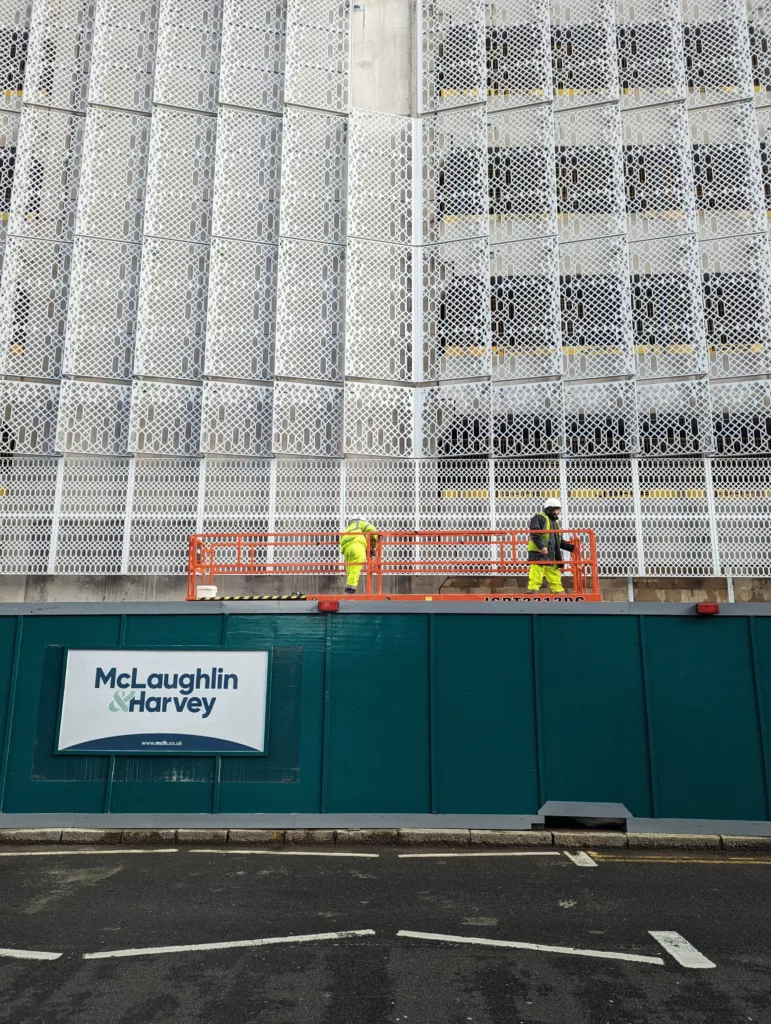 Two workers on an orange lift platform against a white lattice building facade above teal McLaughlin & Harvey construction hoarding.