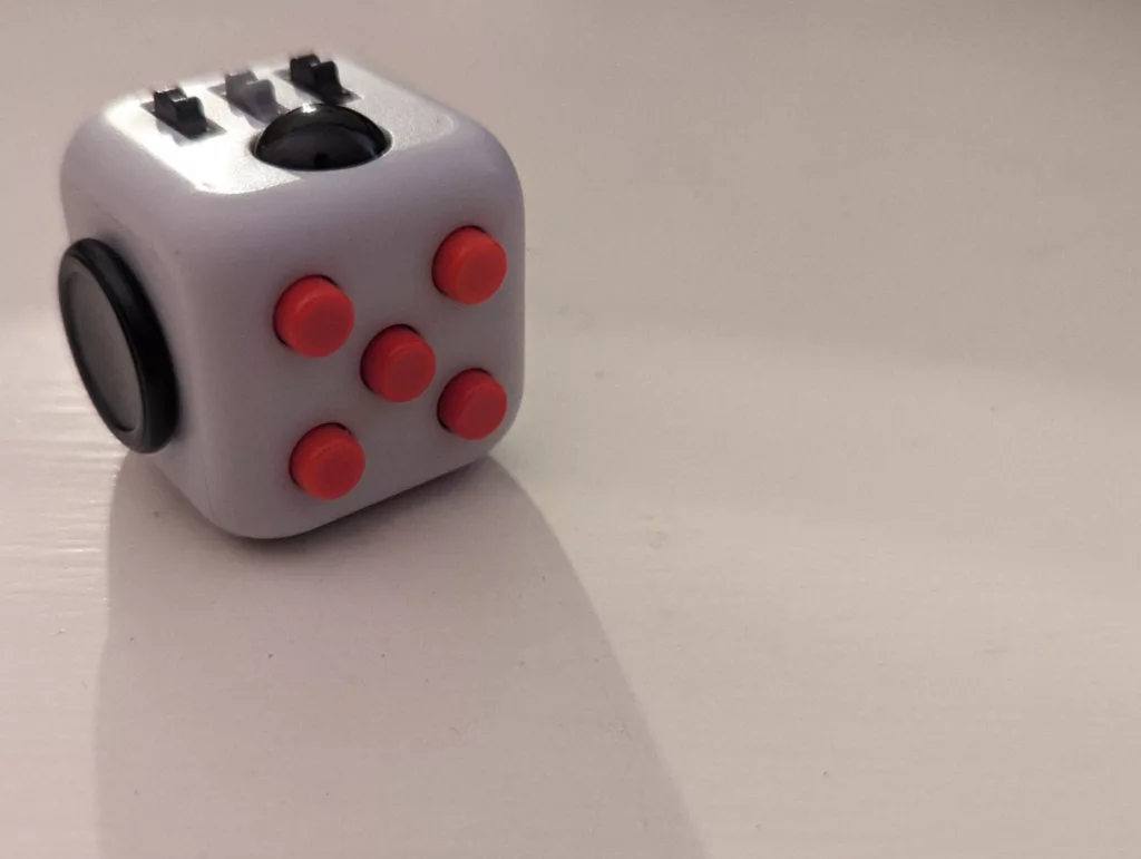 A white fidget cube featuring five red buttons, a black dial, and metal gears, sitting on a plain white surface with a soft shadow.