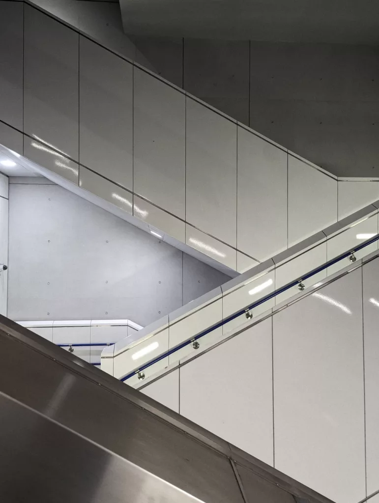 Minimalist architectural view of diagonal white wall panels and a blue-striped handrail in a modern concrete station interior.