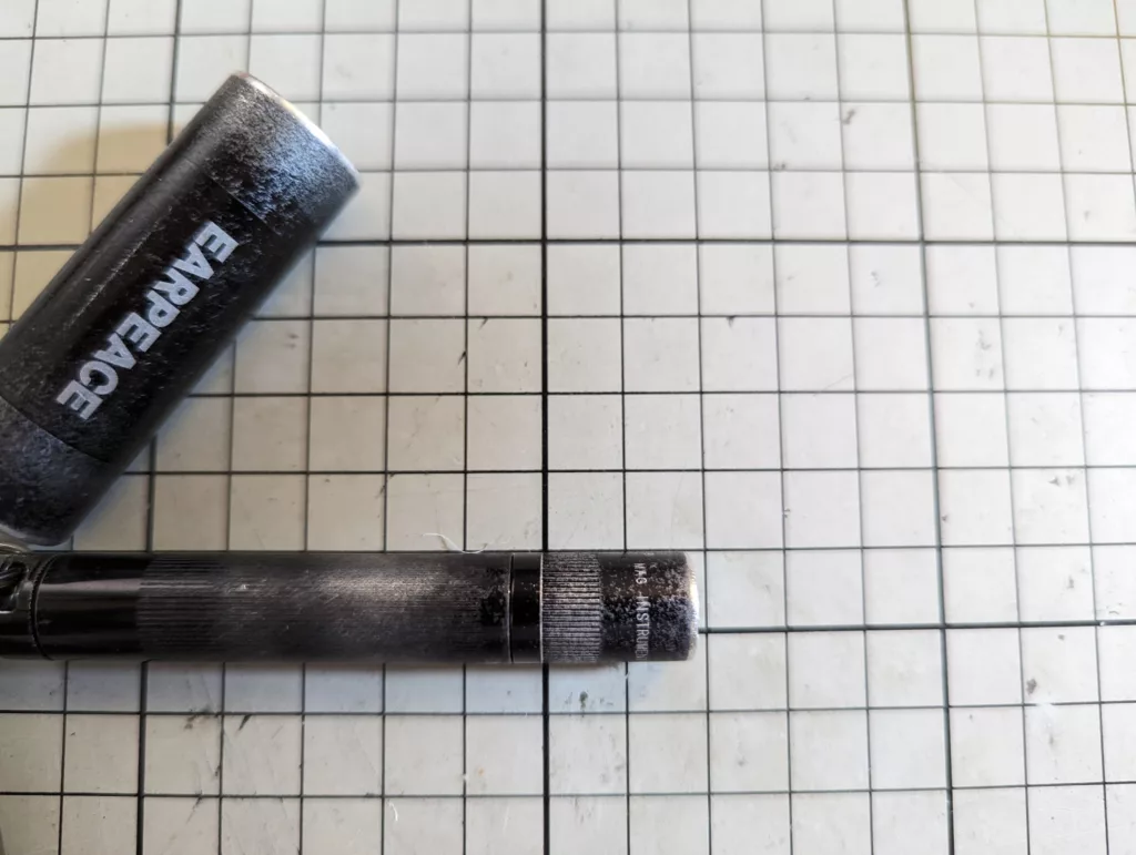 A worn black Earpeace earplug case and a thin black tool with a textured grip on a white gridded cutting mat.