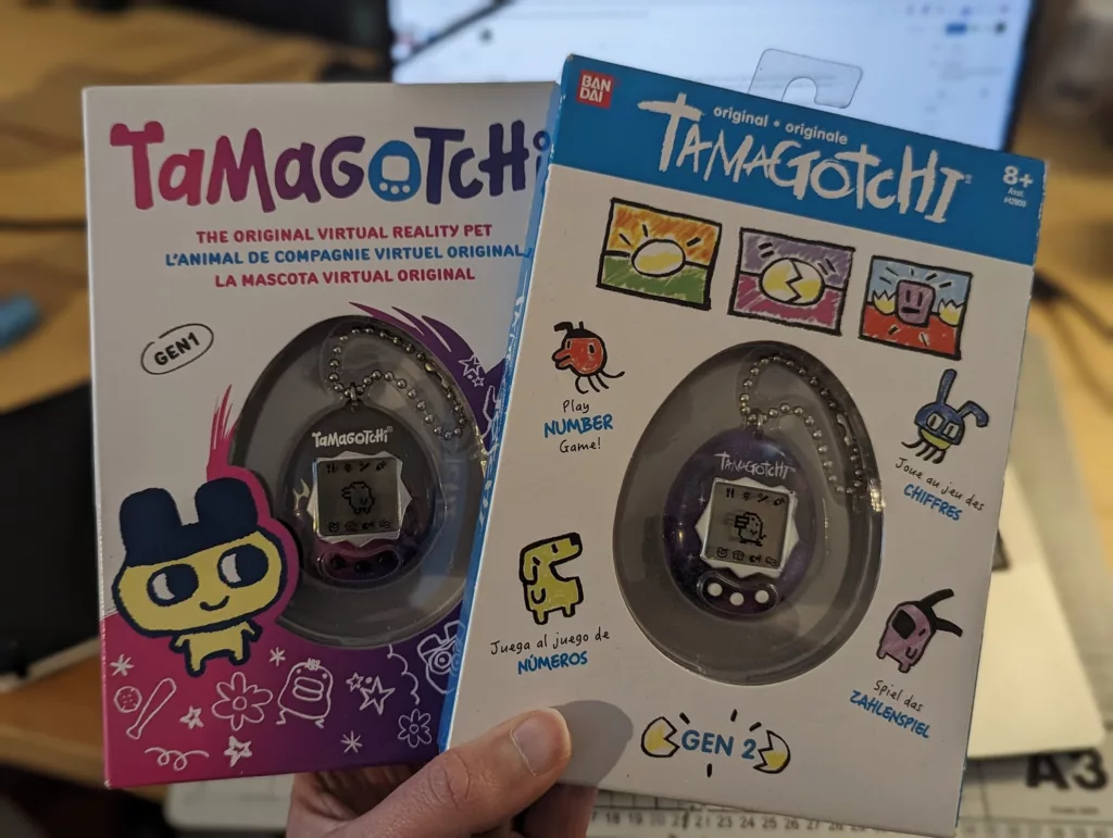 Two boxed original Tamagotchi virtual pets, Gen 1 and Gen 2, held in a hand in front of a computer screen.
