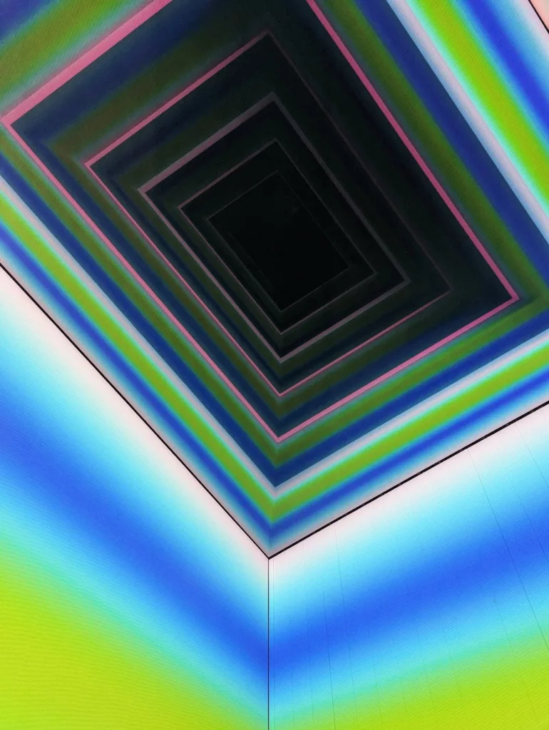 Concentric rectangles in vibrant pink, blue, and green gradients receding into a dark center in a geometric light installation.