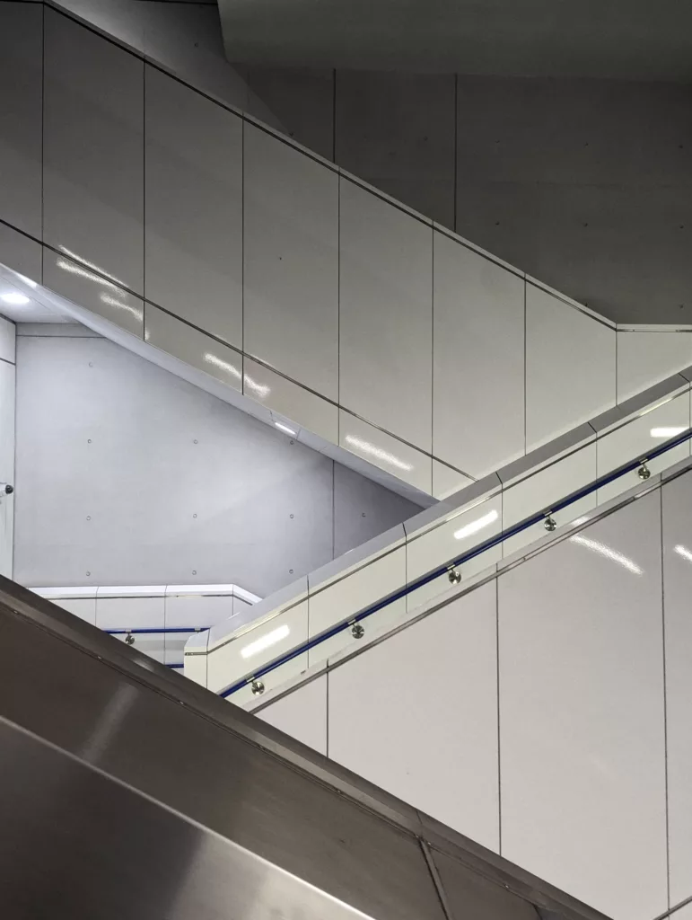 Geometric architectural detail of white glossy panels and a blue handrail forming intersecting diagonal lines against a grey concrete wall.