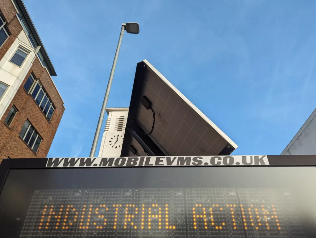A digital road sign displaying INDUSTRIAL ACTION in orange LEDs against a blue sky, with a solar panel and a clock tower in the background.