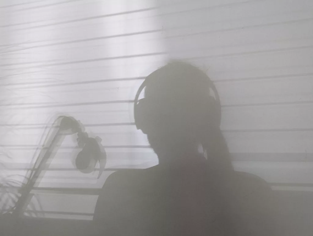 Silhouette of a person wearing headphones next to a microphone, cast on a wall with horizontal light and shadow from window blinds.