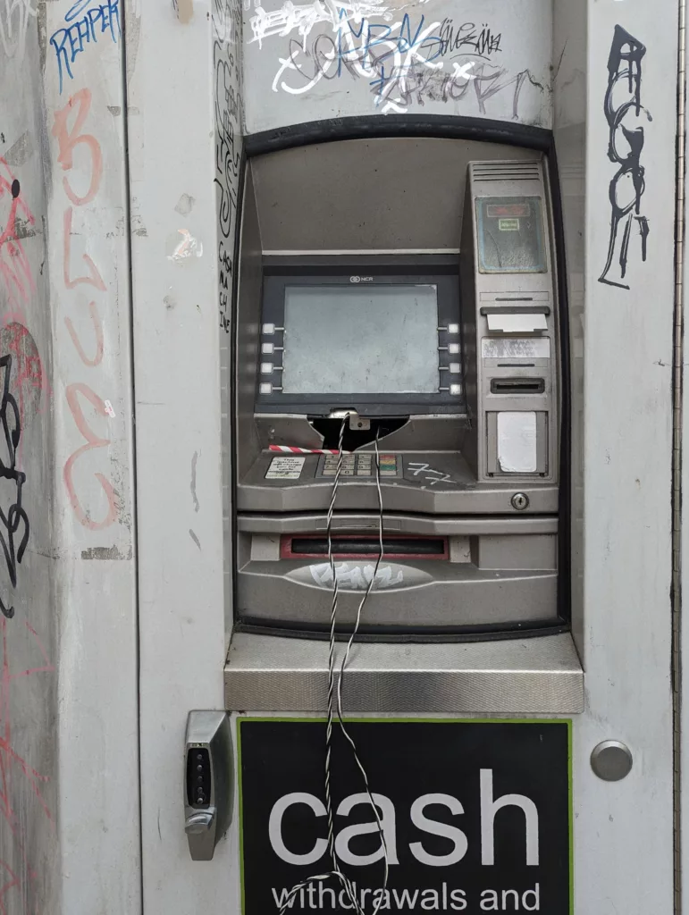 A vandalised cash machine covered in graffiti tags with wires hanging from a smashed hole above the keypad.