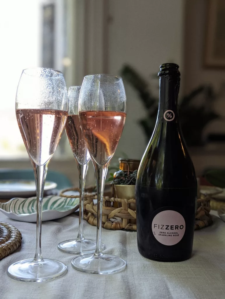Three glasses of sparkling pink rosé beside a bottle of Fizzero non-alcoholic wine on a sunlit table with a white cloth.