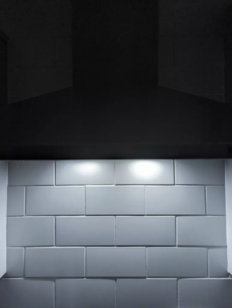 White subway tiles illuminated by two bright overhead lights beneath the dark silhouette of a kitchen extractor hood.