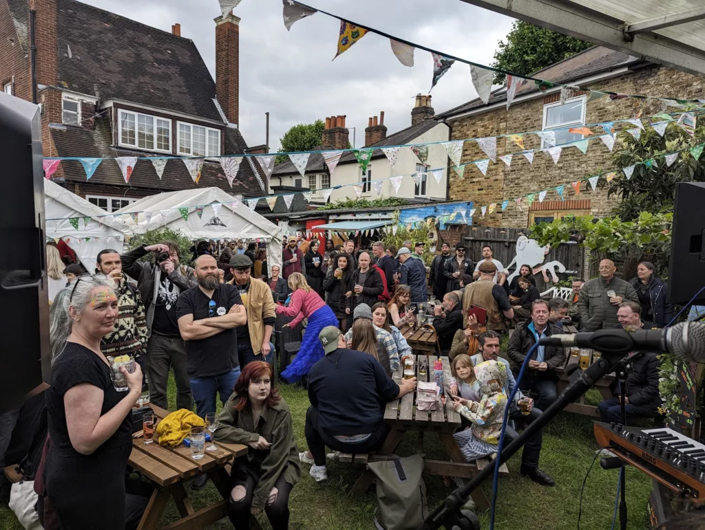 Crowded pub garden with people at picnic tables under festive bunting, viewed from behind a live music microphone and keyboard setup.
