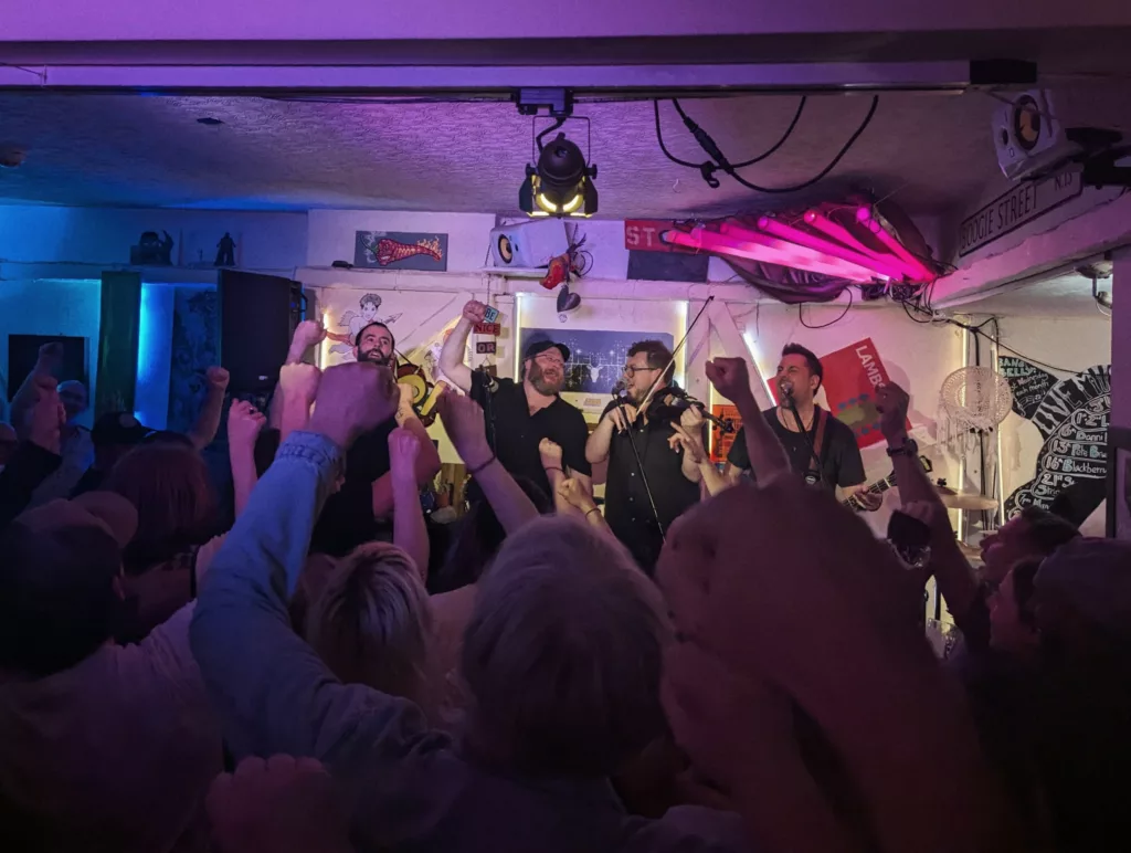A band performs on a small stage under pink lights as a crowd in the foreground cheers with their fists raised in the air.