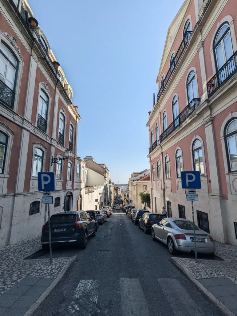 Steep street in Lisbon lined with pink buildings and parked cars, looking downhill toward a distant bridge under a clear blue sky.