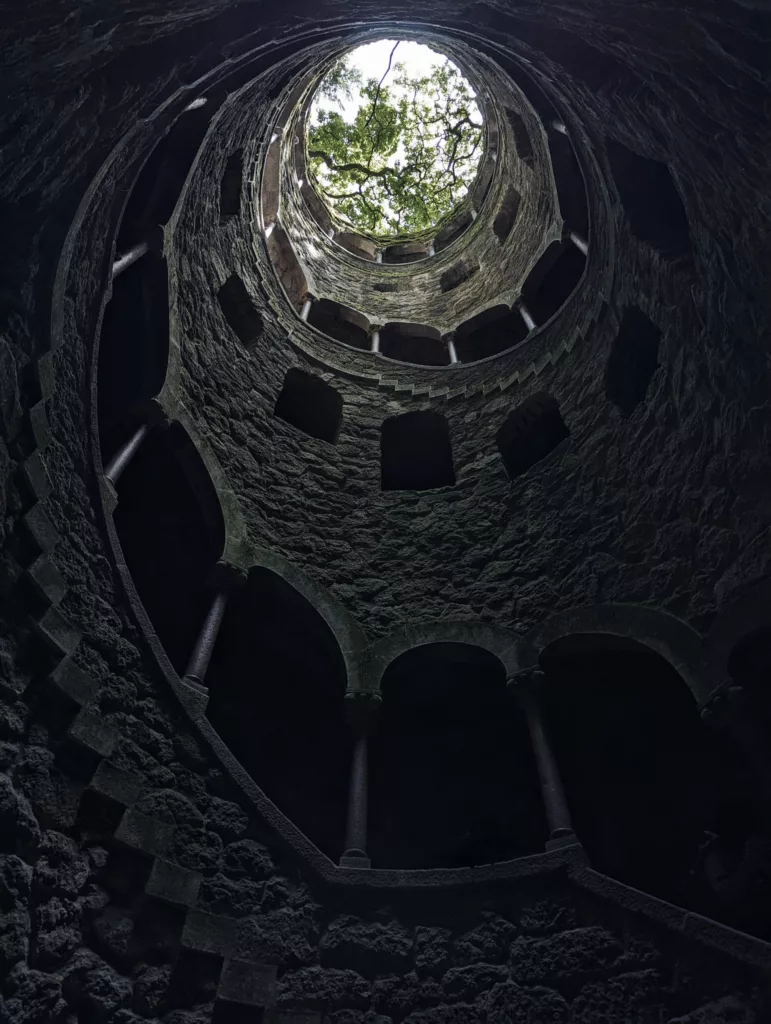 Looking up through the spiral stone arches of the Initiation Well towards a bright circular opening filled with green tree leaves.