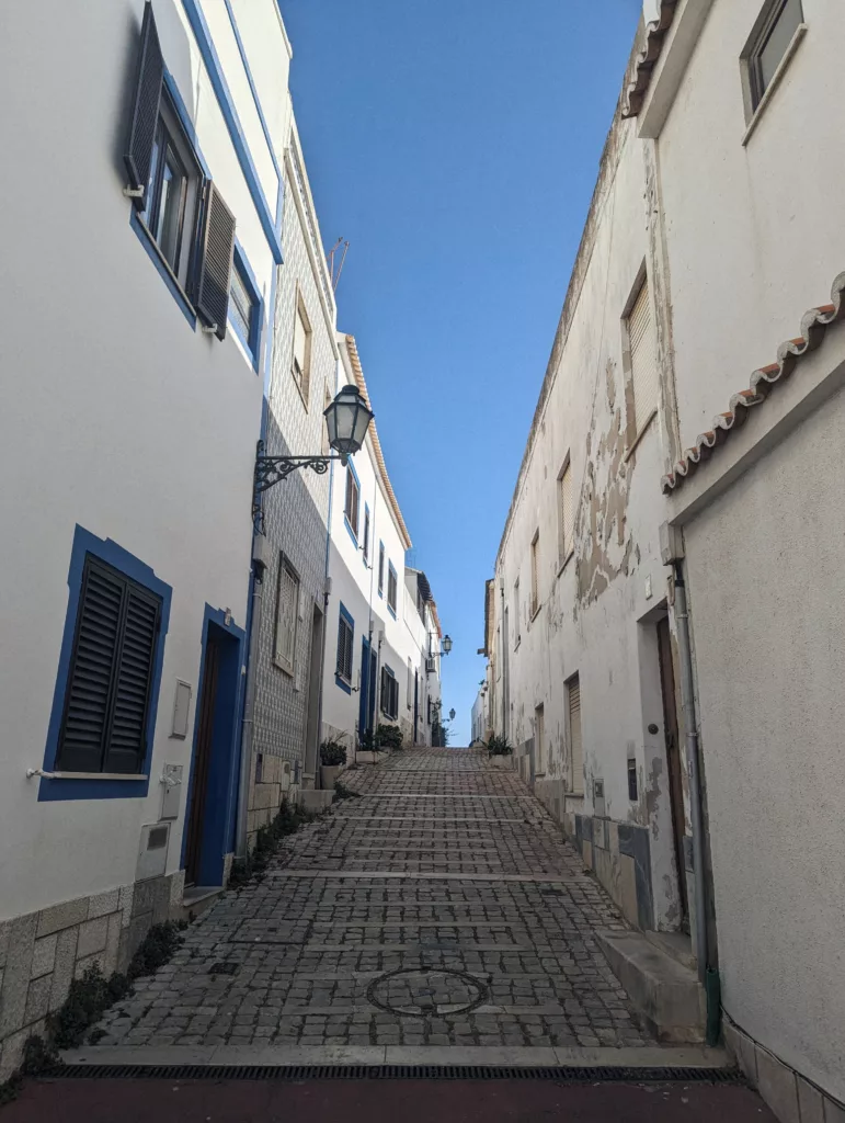 A narrow cobblestone street climbing steeply between white buildings with blue trim and black street lamps under a clear blue sky.