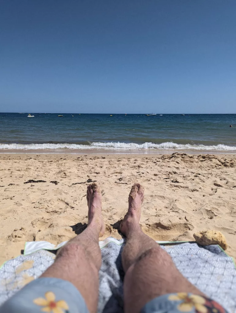 Sandy feet and legs from a first-person view, resting on a patterned towel on a beach facing blue ocean waves and a clear sky.