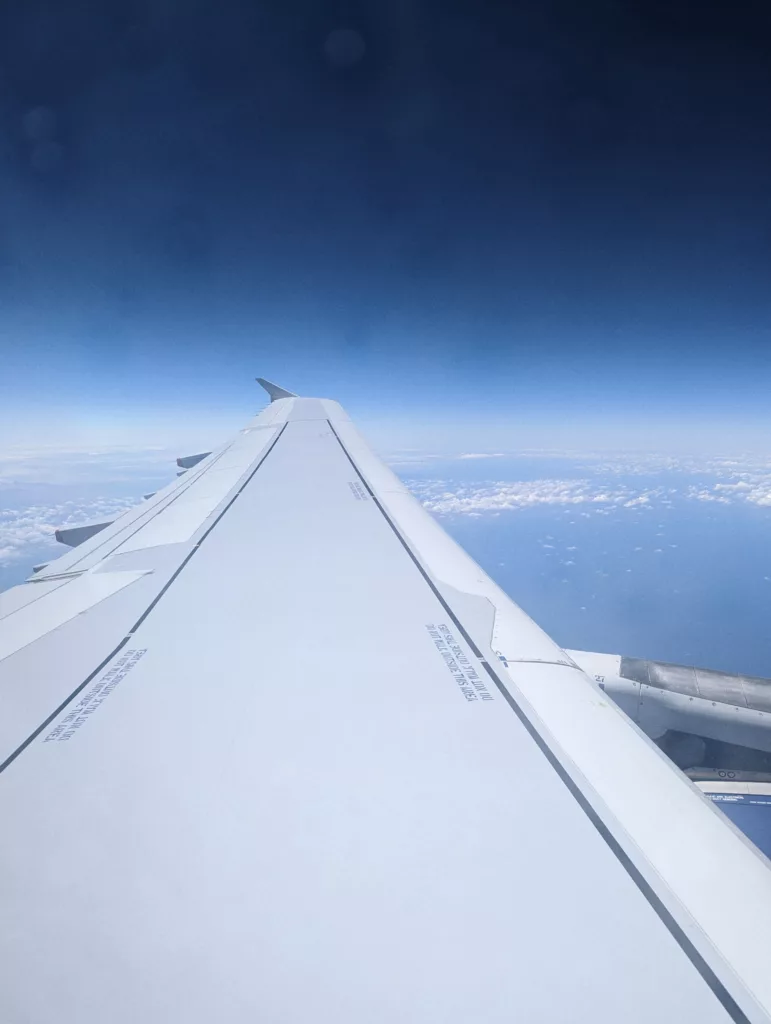 A white airplane wing with safety markings flying over white clouds and a deep blue sky, viewed from a window.