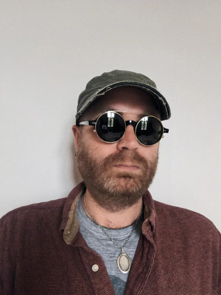 Bearded man wearing round sunglasses, a distressed green baseball cap, and a red corduroy shirt against a white wall.