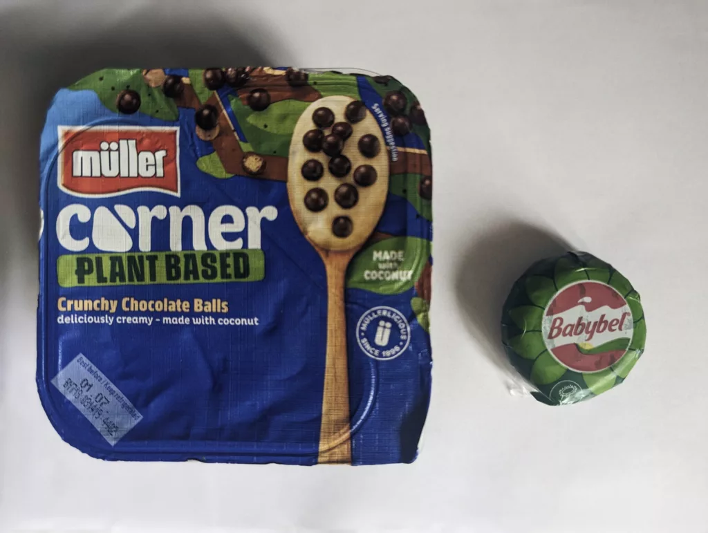 A blue Müller Corner Plant Based yoghurt pot with crunchy chocolate balls beside a green-wrapped Babybel snack on a white background.