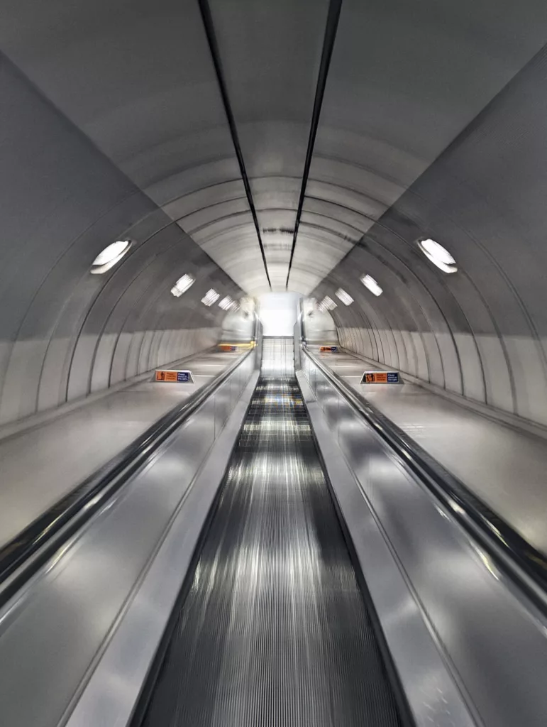 Long exposure of a symmetrical escalator in a curved underground tunnel with rows of glowing lights leading into the distance.