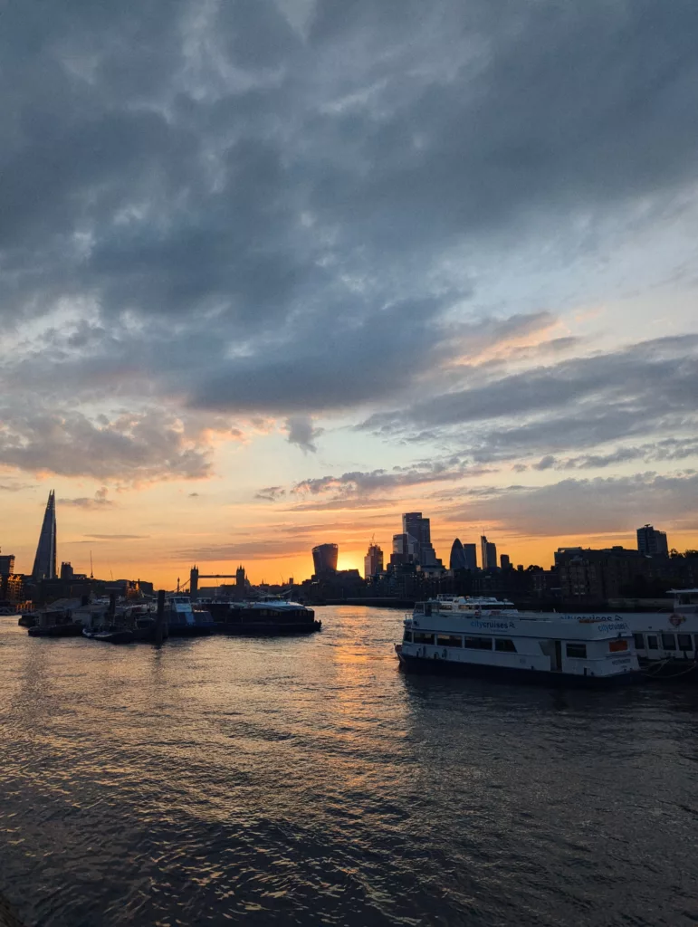 Sunset over the River Thames with tour boats and the London skyline silhouetted against a dramatic orange and grey cloudy sky.