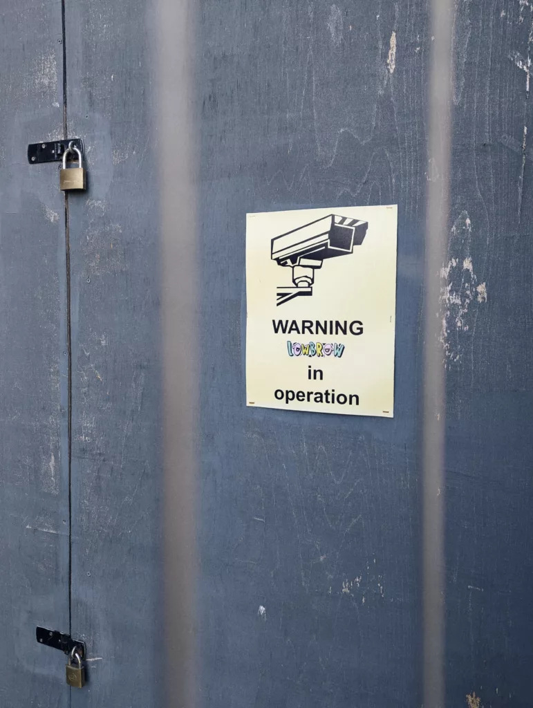 A yellow sign on a blue wooden door reads "WARNING LOWBROW in operation" below a CCTV icon. Two brass padlocks are visible on the left.