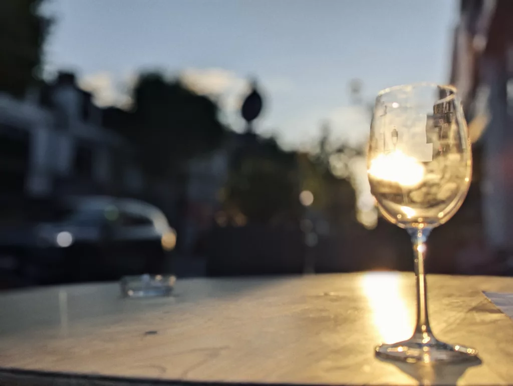 Golden hour sunlight glows through a wine glass on a wooden cafe table, with a blurred street scene in the background.