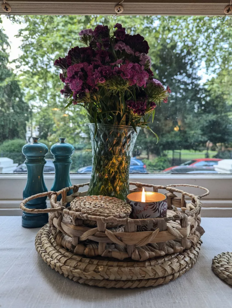 A bouquet of purple flowers in a glass vase, a lit candle, and teal grinders on a woven tray by a window overlooking leafy green trees.