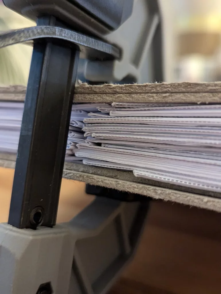 A stack of paper signatures sandwiched between grey boards and held firmly in a black and grey bar clamp.