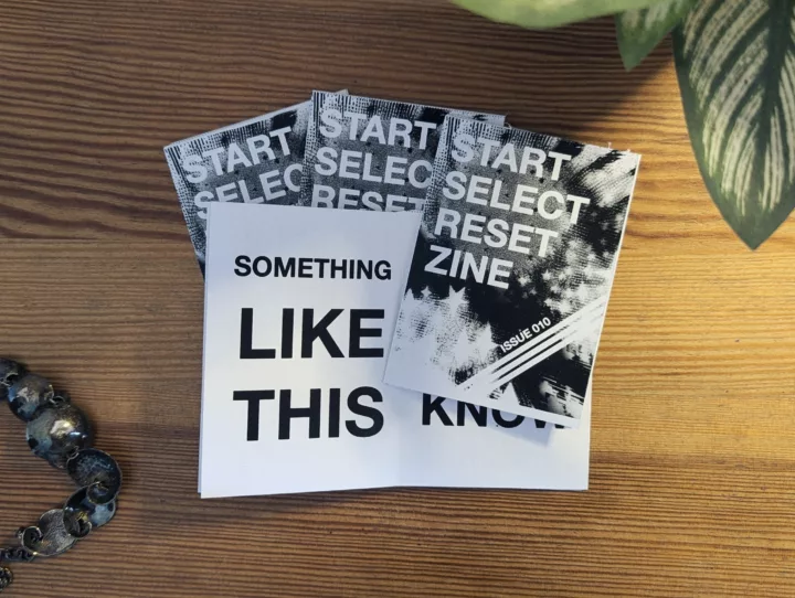 Copies of Start Select Reset Zine on a wooden surface, one open to a page reading SOMETHING LIKE THIS in bold capital letters.