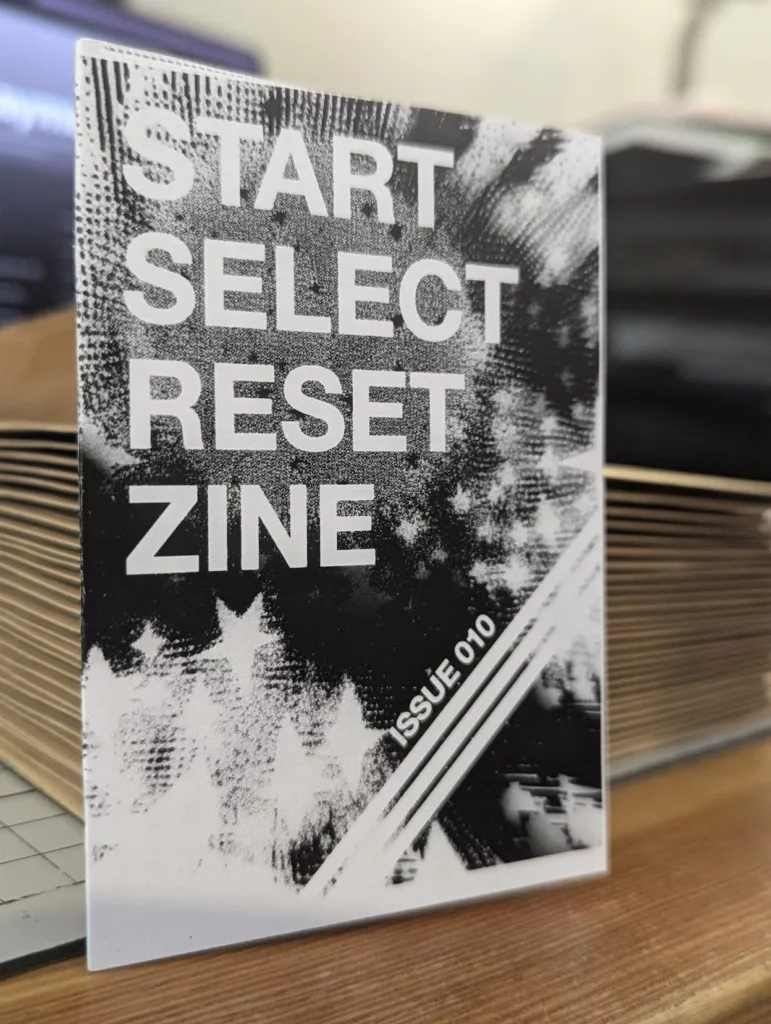 Black and white grainy cover for START SELECT RESET ZINE ISSUE 010 with a star pattern, leaning against a stack of booklets on a desk.